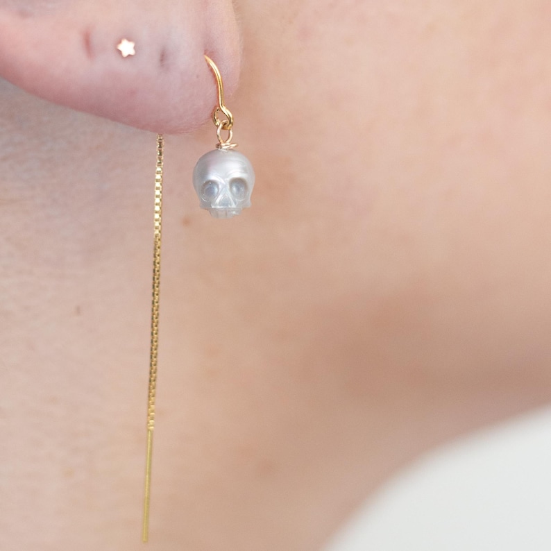 May include: A gold-colored earring with a small, gray skull charm. The earring has a long, thin chain that hangs down. A small star-shaped stud earring is also visible.