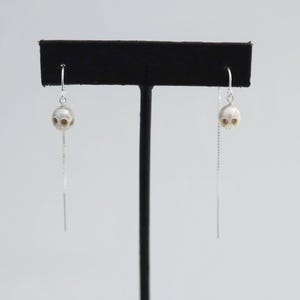 May include: A pair of silver threader earrings featuring small, carved white skull beads. Each earring consists of a delicate silver chain that passes through the ear, with a single skull bead suspended from a silver hook at the front.