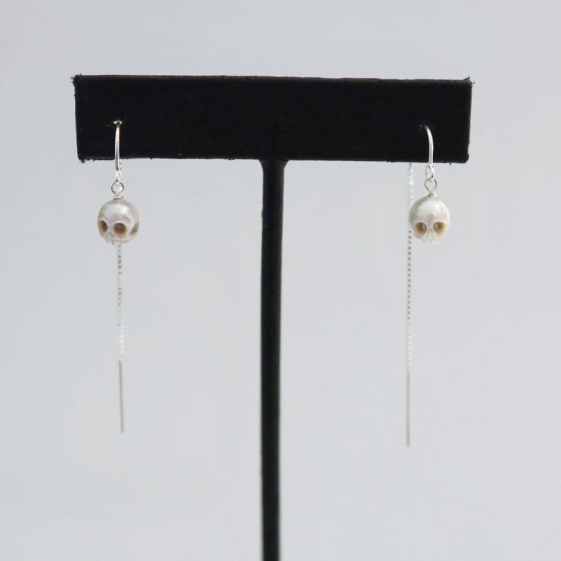 May include: A pair of silver threader earrings featuring small, carved white skull beads. Each earring consists of a delicate silver chain that passes through the ear, with a single skull bead suspended from a silver hook at the front.