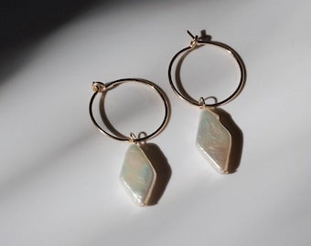 May include: A pair of gold hoop earrings with a pearlescent, diamond-shaped charm. The earrings are displayed on a white surface, with shadows adding depth. The charms have a subtle iridescent quality.
