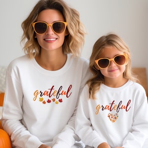 Pode incluir: Duas blusas de moletom brancas com a palavra "grateful" impressa nelas. Uma blusa de moletom tem uma fileira de pequenas folhas de outono abaixo da palavra, e a outra tem um desenho em forma de coração feito de folhas de outono.