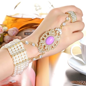May include: A gold bracelet with a large purple rhinestone and smaller rhinestones. The bracelet is connected to a ring with rhinestones. The bracelet is on a person's wrist.