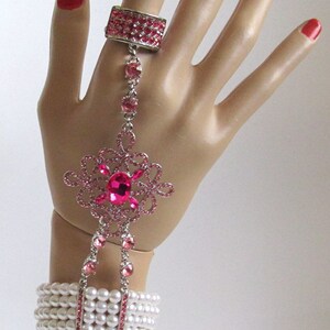May include: A pink rhinestone bracelet with a large pink rhinestone flower charm and a matching ring. The bracelet is made of white pearls and has a silver chain connecting the ring to the bracelet.