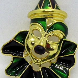 May include: A gold-tone clown mask brooch with green and black enamel. The mask has a wide smile and a black nose. The clown is wearing a green and purple hat.