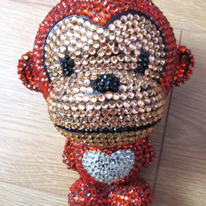 May include: A red, orange, and black monkey figurine covered in rhinestones. The monkey has a heart-shaped design on its chest.
