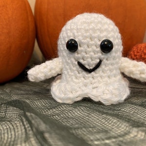 May include: A handmade white crochet ghost with black button eyes and a stitched black smile. The ghost is positioned in front of two orange pumpkins and a small crocheted pumpkin, creating a Halloween theme.
