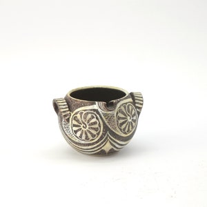 Puede incluir: Una pequeña olla de cerámica marrón con diseños geométricos blancos. La olla tiene dos pequeñas asas a los lados.