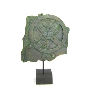 Puede incluir: Un engranaje de metal verde con un diseño en forma de cruz, montado en un soporte negro. El engranaje es circular con un cubo central y cuatro radios.