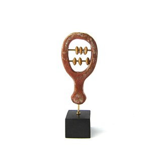 May include: A decorative wooden abacus sculpture. The sculpture is reddish-brown with a distressed finish, featuring two rows of golden beads. It is mounted on a black square base, connected by a thin gold rod. The background is white.