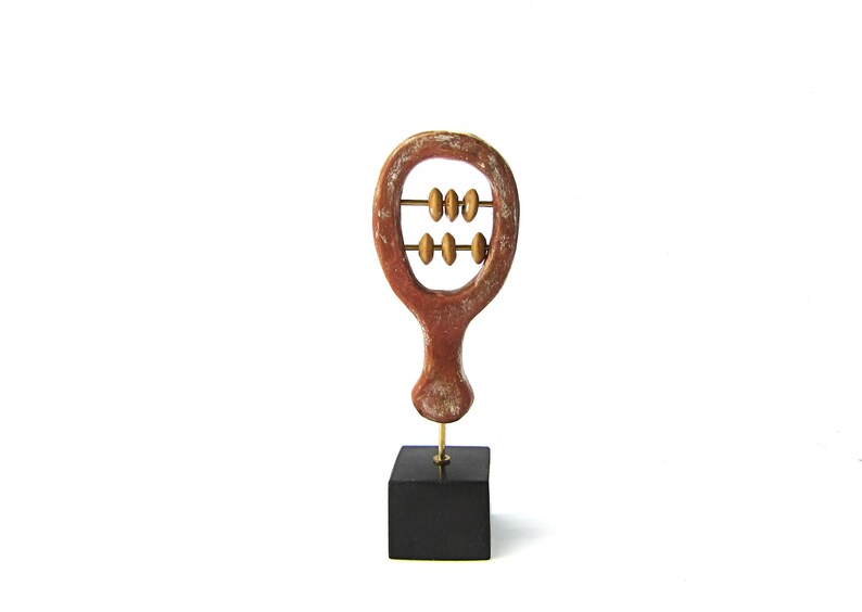 May include: A decorative wooden abacus sculpture. The sculpture is reddish-brown with a distressed finish, featuring two rows of golden beads. It is mounted on a black square base, connected by a thin gold rod. The background is white.