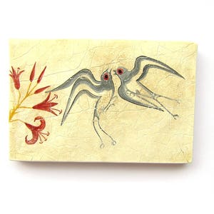 May include: A rectangular, off-white decorative piece with a painted design. Two stylized birds with gray wings and red eyes face each other. Red flowers and yellow stems are on the left side. The background has a textured appearance.