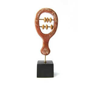 May include: A handcrafted, abstract sculpture resembling an abacus. The reddish-brown wooden frame holds two rows of golden beads. It is mounted on a small, black square base, creating a minimalist aesthetic.