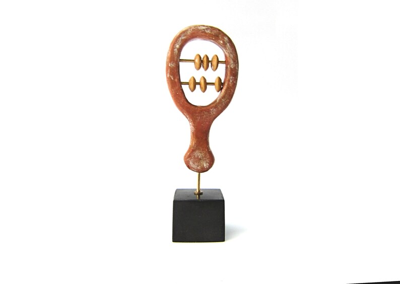 May include: A handcrafted, abstract sculpture resembling an abacus. The reddish-brown wooden frame holds two rows of golden beads. It is mounted on a small, black square base, creating a minimalist aesthetic.