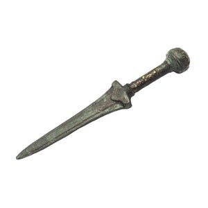 May include: An antique bronze dagger with a rounded pommel and a long, pointed blade. The handle features intricate gold-colored detailing. The dagger is displayed against a white background, highlighting its aged appearance and historical significance.