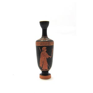 Puede incluir: Jarr&oacute;n de cer&aacute;mica negro y rojo con un dise&ntilde;o griego que representa una figura sosteniendo una lanza. El jarr&oacute;n tiene un cuello estrecho y una base redonda.