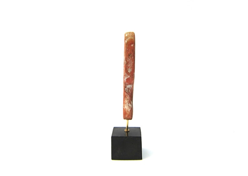 May include: A vertical, weathered, reddish-brown wooden object with a gold-colored metal pin, mounted on a small, black, square base. The wood has a textured surface with lighter patches. The object stands against a white background.