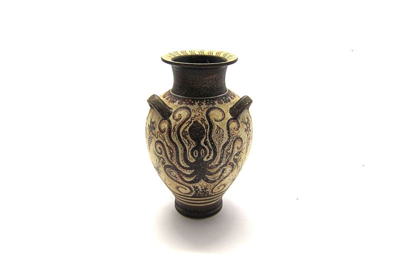 May include: A small, cream-coloured ceramic vase with a brown octopus design. The vase has two handles and a narrow neck.