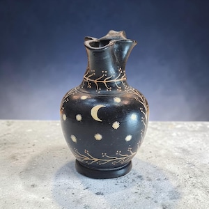 May include: A black ceramic pitcher with a unique, curved spout. The pitcher is decorated with a crescent moon, stars, and tree branch designs in a contrasting light color. The base is a dark, circular shape.
