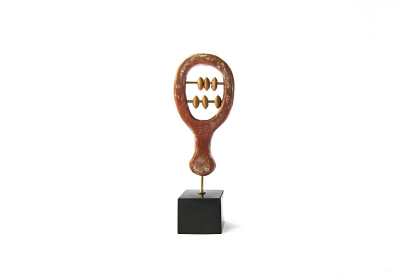 May include: A decorative abacus sculpture with a reddish-brown frame and golden beads. The abacus is mounted on a brass rod and a black square base. The background is plain white.