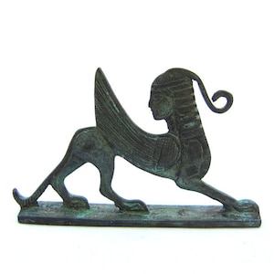 May include: A green bronze statue of a winged sphinx with a human head and a lion's body. The sphinx is depicted in a crouching position with its wings spread out behind it.
