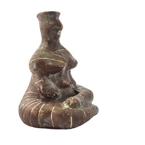 May include: A brown ceramic figurine of a seated woman with stylized features and incised lines. The figurine has a rounded, bulbous shape and a simple, abstract design.