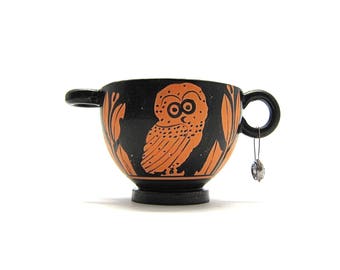 May include: A black and orange ceramic cup with a handle and an owl design. A silver earring with a clear stone hangs from the handle. The cup has a pedestal base against a white background.