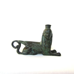 Op de afbeelding: Een groen bronzen beeld van een gevleugelde sfinx met een menselijk hoofd en een leeuwenlichaam.