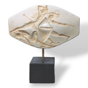 Pode incluir: Uma escultura em relevo de cor creme que retrata uma cena de batalha com figuras empunhando armas. A escultura é montada em uma haste de metal dourado e uma base quadrada preta. A obra de arte é provavelmente uma peça decorativa.
