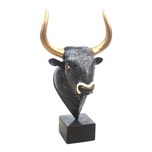 Op de afbeelding: Zwarte stierenkop sculptuur met gouden hoorns op een zwarte basis.