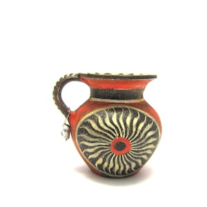 Puede incluir: Pequeña jarra decorativa de cerámica, roja con detalles en negro y blanco y un diseño de sol. El asa es texturizada y tiene un pequeño adorno colgando. Hecha a mano, es ideal para la decoración.