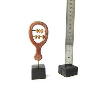May include: A small, decorative abacus sculpture with a reddish-brown finish, mounted on a black square base. The abacus has three rows of beads. A metal ruler is next to the abacus, showing measurements in inches.