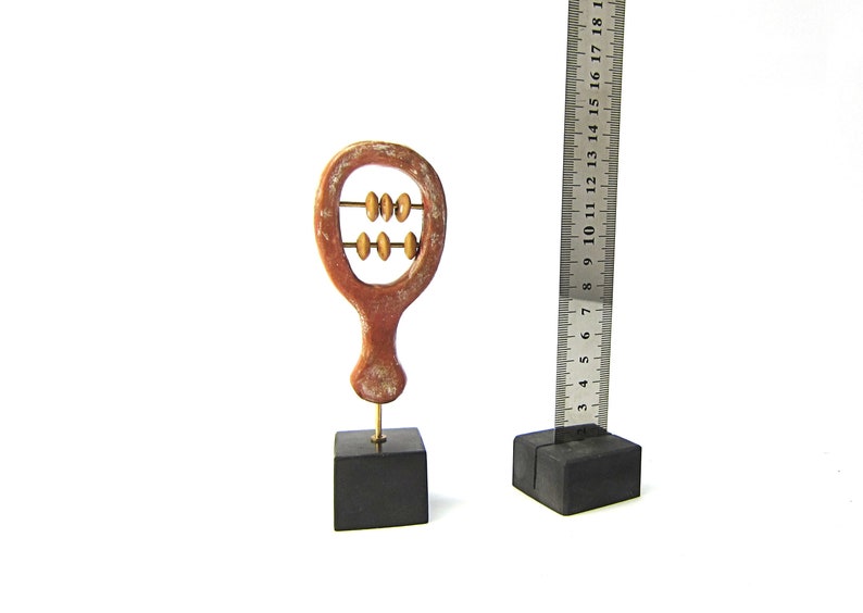 May include: A small, decorative abacus sculpture with a reddish-brown finish, mounted on a black square base. The abacus has three rows of beads. A metal ruler is next to the abacus, showing measurements in inches.