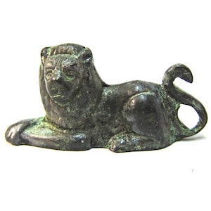 Könnte beinhalten: Eine kleine Bronzeskulptur eines liegenden Löwen mit einem eingerollten Schwanz. Die Skulptur ist detailliert mit einer strukturierten Oberfläche und hat eine grüne Patina.