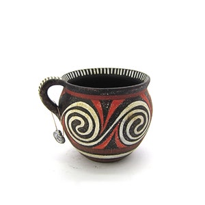 Puede incluir: Una pequeña olla de cerámica marrón con asa. La olla está decorada con patrones geométricos rojos, blancos y negros, incluyendo espirales.