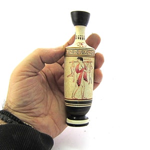 Puede incluir: Un pequeño jarrón de cerámica con la parte superior negra y la base blanca. El jarrón está decorado con una imagen roja y blanca de una persona que lleva dos animales en un palo. El jarrón tiene un diseño de clave griega alrededor de la parte superior.