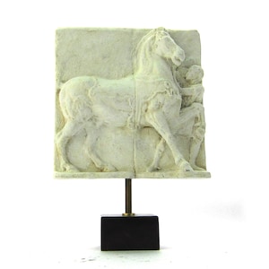 Op de afbeelding: Witte gipsen reliëfsculptuur van een paard en een persoon op een zwarte standaard. Het beeld is een reproductie van een klassiek Grieks kunstwerk.