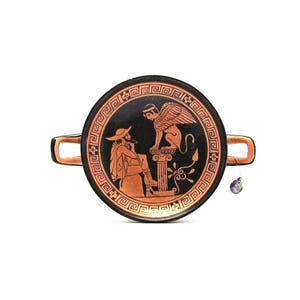 Puede incluir: Un plato de cerámica negro y rojo con un diseño griego que presenta una criatura alada y una persona. El plato tiene un asa a cada lado.