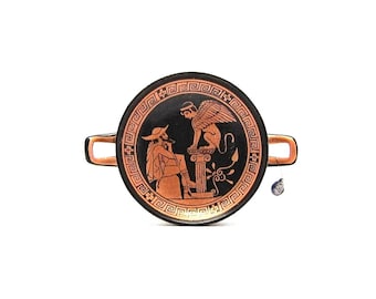 May include: A black and red ceramic plate with a Greek design featuring a winged creature and a person. The plate has a handle on each side.