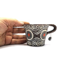 Puede incluir: Una taza pequeña de cerámica marrón con un diseño geométrico blanco y negro. La taza tiene un asa y dos círculos rojos en la parte delantera.