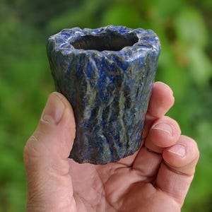 May include: A handmade, small ceramic cup with a textured, bark-like exterior. The cup is primarily a deep blue color with hints of green and brown, and has a rough, uneven rim. The cup is held in a hand against a blurred green background.