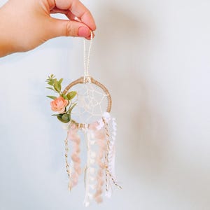 K&ouml;nnte beinhalten: Ein kleiner Traumf&auml;nger mit einem wei&szlig;en und braunen Makrameemuster, rosa und wei&szlig;en Federn und einer einzelnen rosa Blume.