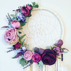 May include: A white dream catcher with a circular frame and a web-like design. The frame is decorated with a garland of purple, pink, and burgundy flowers.