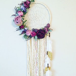 May include: A white dream catcher with a floral wreath made of purple, pink, and green flowers. The dream catcher has white and gold feathers hanging from the bottom.