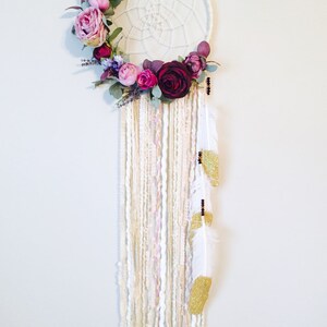 May include: A white dream catcher with a floral wreath made of pink, purple, and burgundy flowers. The dream catcher has white and gold yarn strands and white feathers with gold glitter accents.
