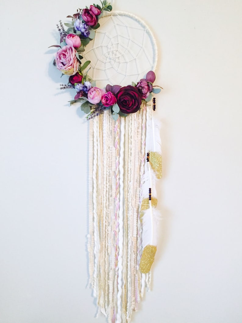 May include: A white dream catcher with a floral wreath made of pink, purple, and burgundy flowers. The dream catcher has white and gold yarn strands and white feathers with gold glitter accents.