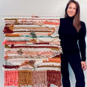 May include: A large, colorful wall hanging made of woven yarn with a variety of textures and colors, including pink, orange, brown, and white. The hanging has a fringe border and is displayed on a white wall.