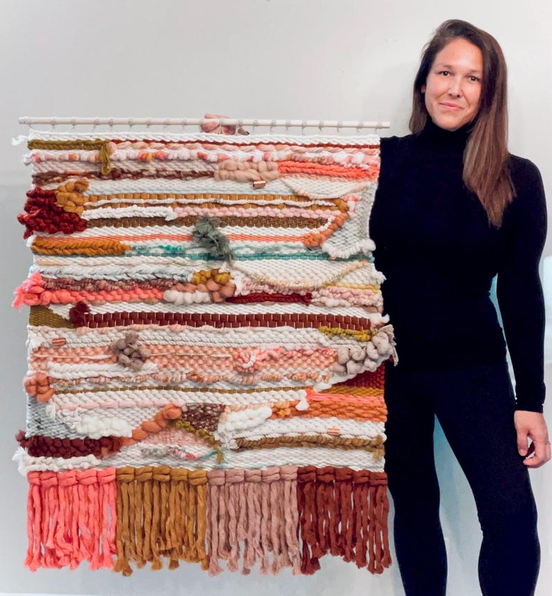 May include: A large, colorful wall hanging made of woven yarn with a variety of textures and colors, including pink, orange, brown, and white. The hanging has a fringe border and is displayed on a white wall.