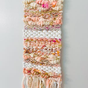 May include: A woven wall hanging with a pink, white, and gold color scheme. The hanging features a variety of textures and patterns, including pom-poms, fringe, and sequins.