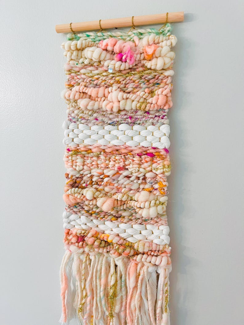 May include: A woven wall hanging with a pink, white, and gold color scheme. The hanging features a variety of textures and patterns, including pom-poms, fringe, and sequins.