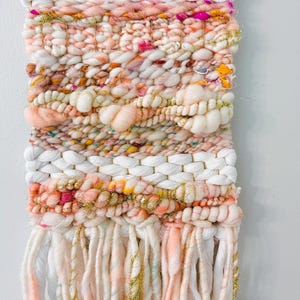 May include: A pink, white, and gold woven wall hanging with fringe. The wall hanging is made with a variety of textures and colors, including fluffy yarn, sparkly thread, and woven strands.